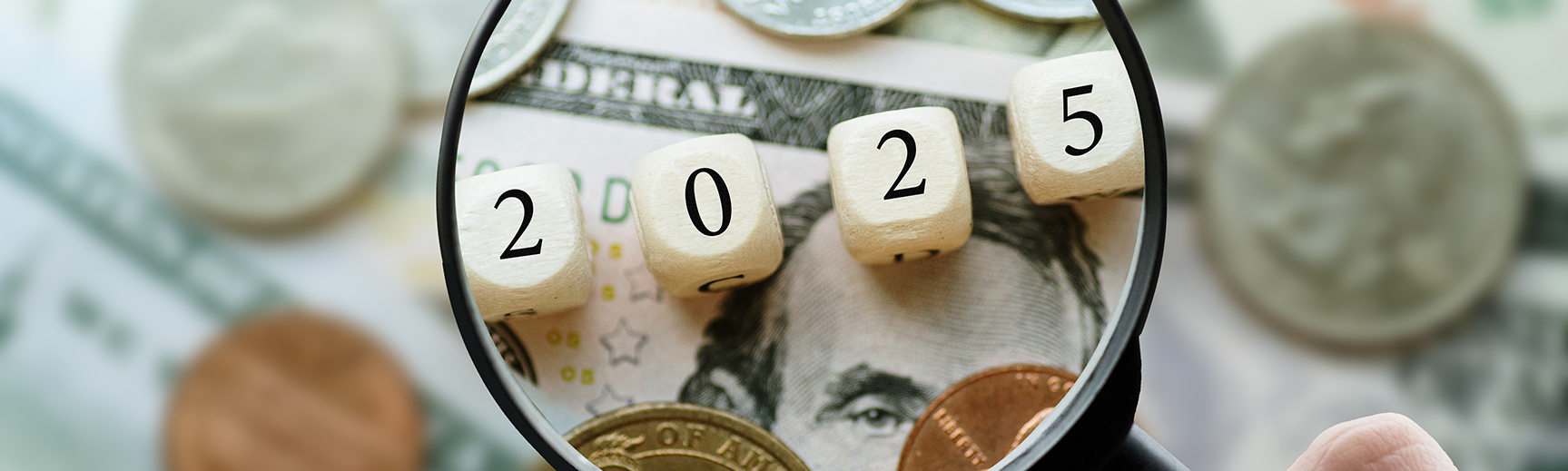 A magnifying glass is sitting on a background of cash, including a five-dollar bill and some change. The magnifying glass is showing four die spelling out "2025."