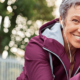 An older woman with short gray hair and wearing a purple hooded jacket puts her foot high on a wall to stretch her muscles while smiling at the camera