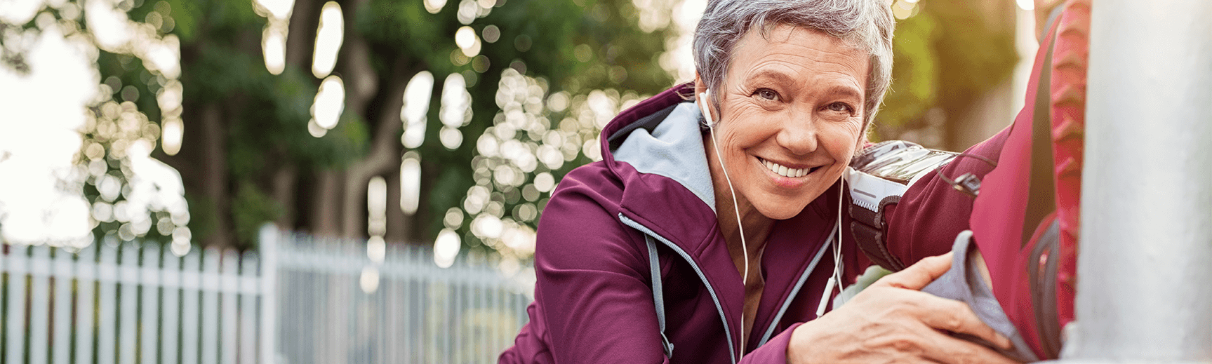 An older woman with short gray hair and wearing a purple hooded jacket puts her foot high on a wall to stretch her muscles while smiling at the camera