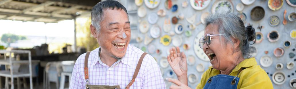 An older couple sitting in front of a wall of painted pottery shares a laugh. The man has paint on his nose.