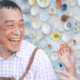 An older couple sitting in front of a wall of painted pottery shares a laugh. The man has paint on his nose.